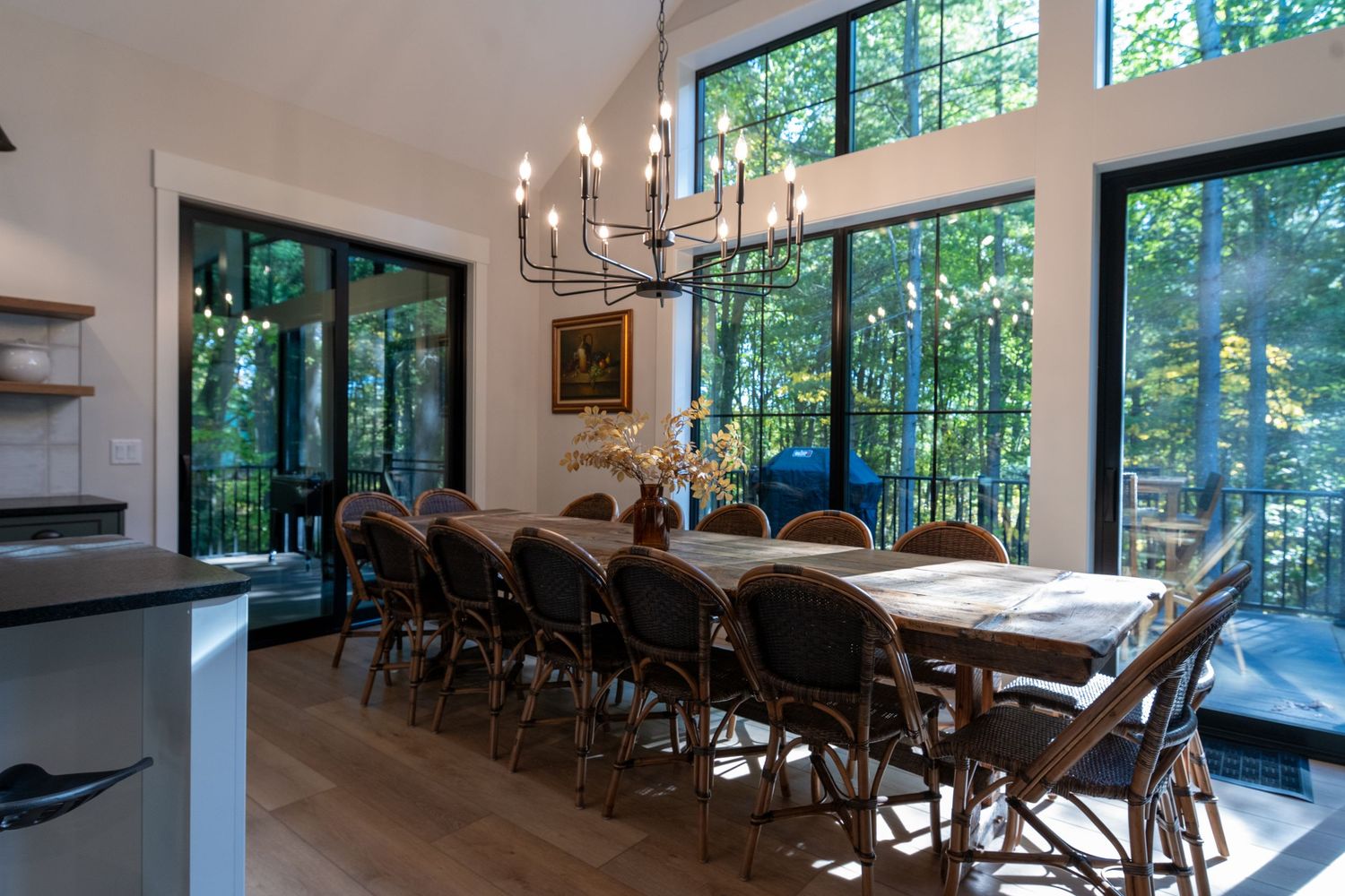 Elegant dining room with large windows overlooking forest, long wooden table, wicker chairs, and chandelier.