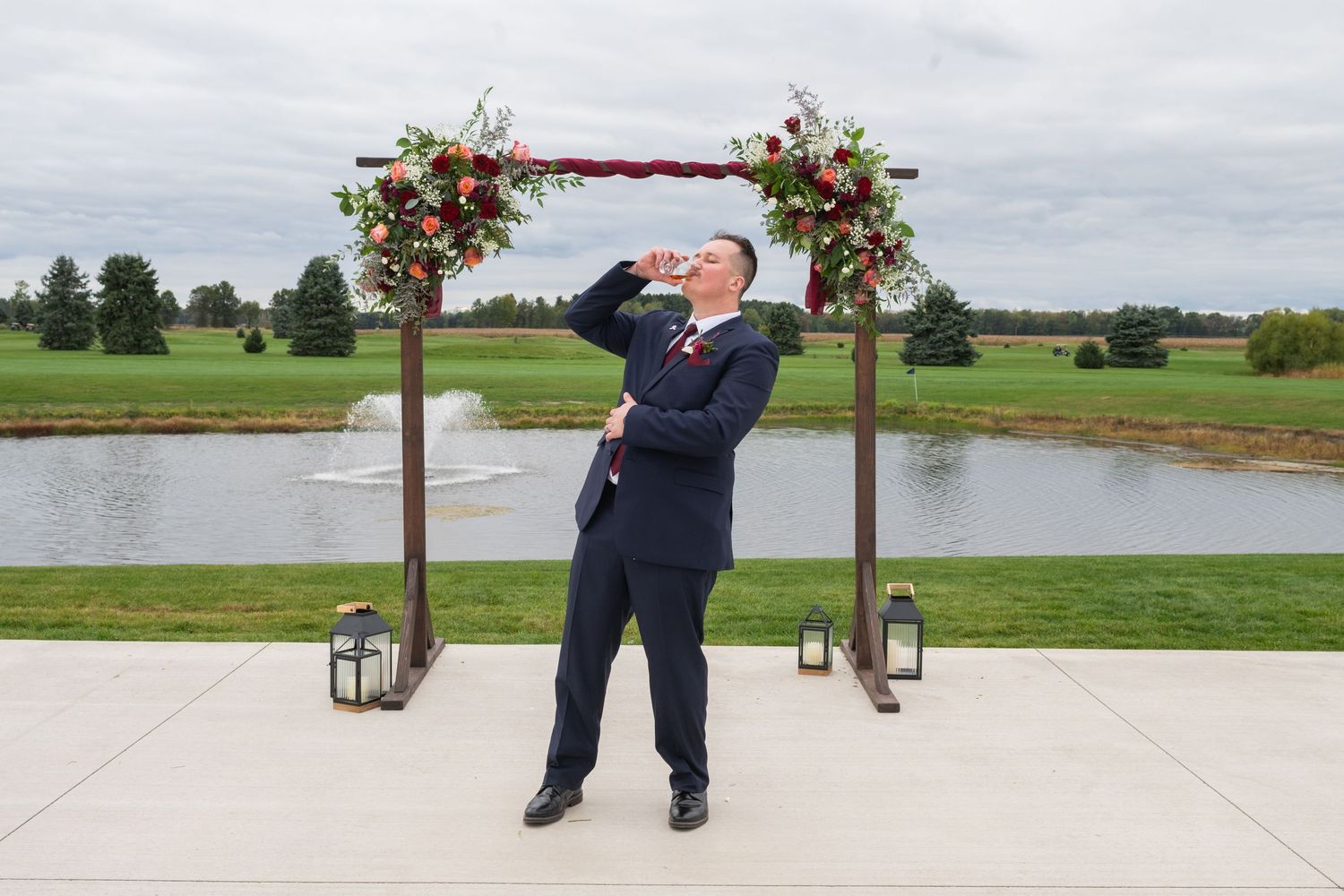 Groom in navy suit celebrating under a floral wedding arch with a scenic pond and green landscape backdrop.