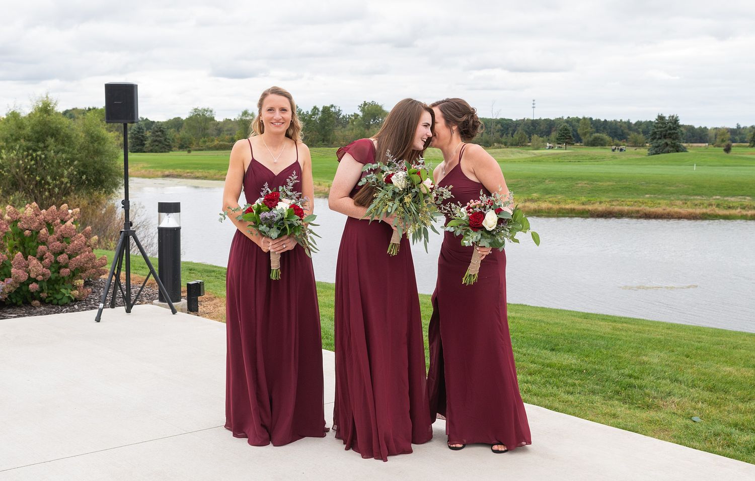 Three bridesmaids in burgundy gowns hold floral bouquets outdoors near a scenic pond.