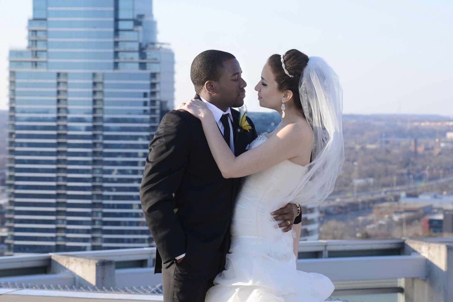 Wedding couple embraces on rooftop with modern city skyline in background.