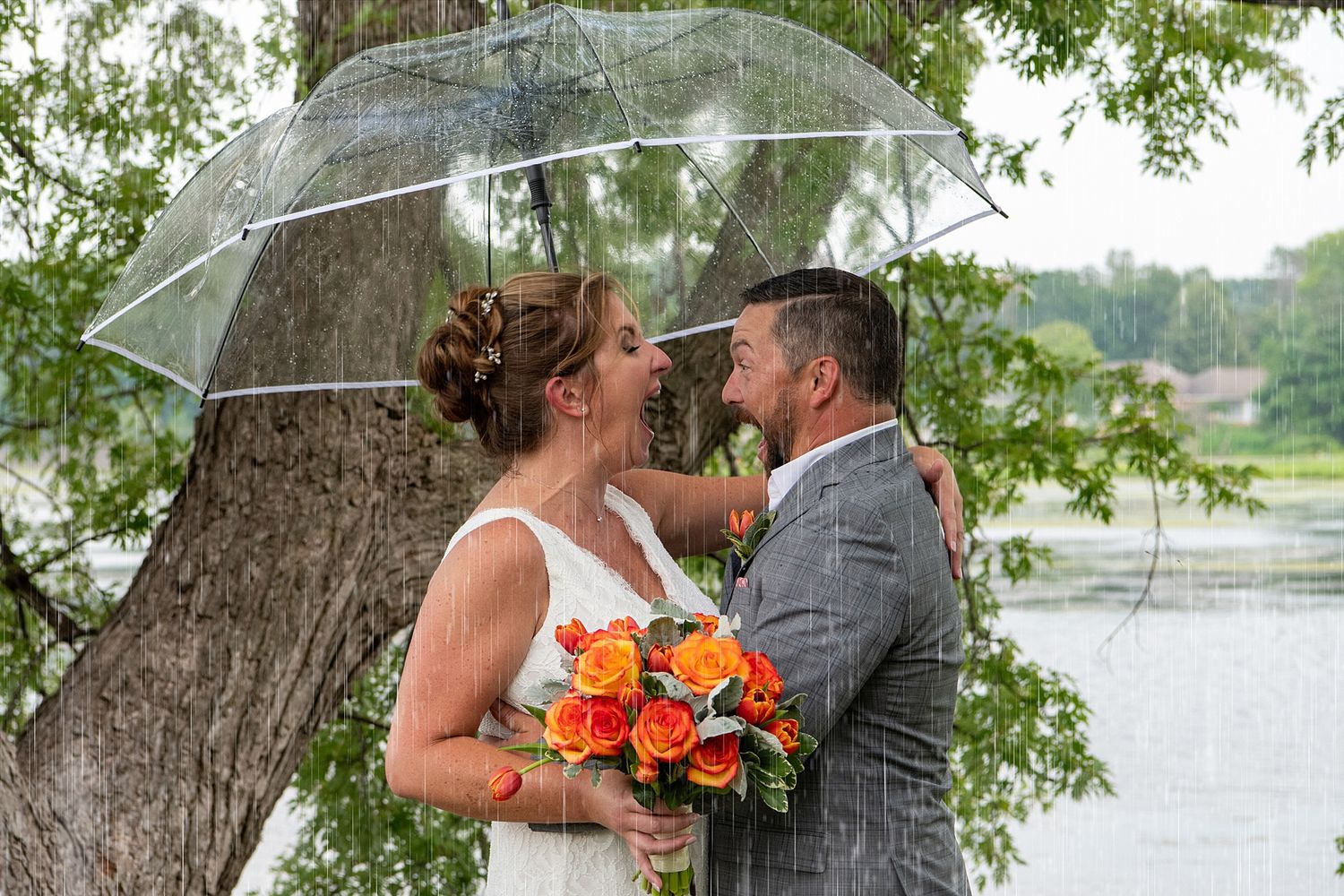 Wedding couple embraces under large tree branches near waterfront.