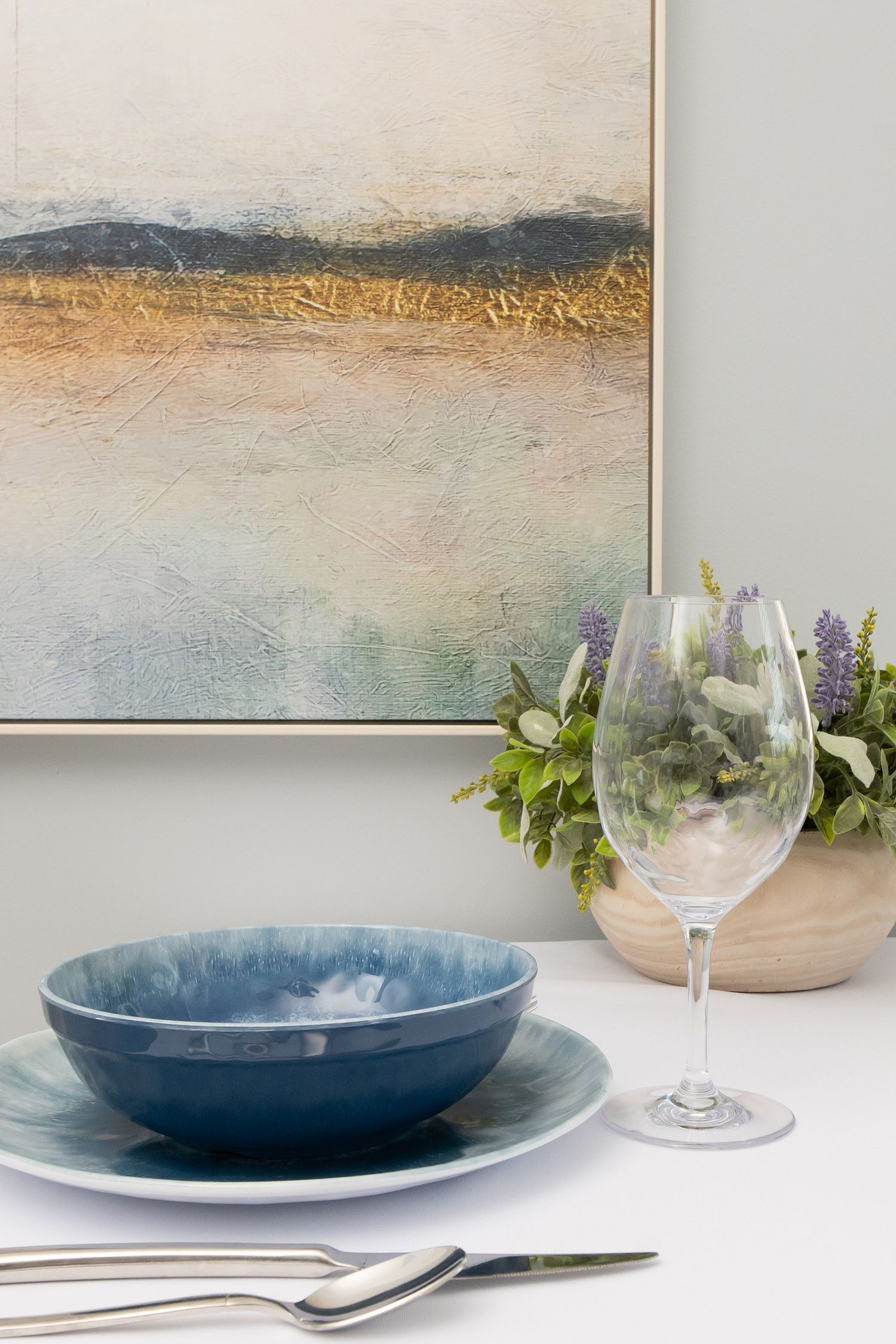 Blue ceramic bowl and plate set with wine glass, silverware, and floral vase beside abstract landscape painting.