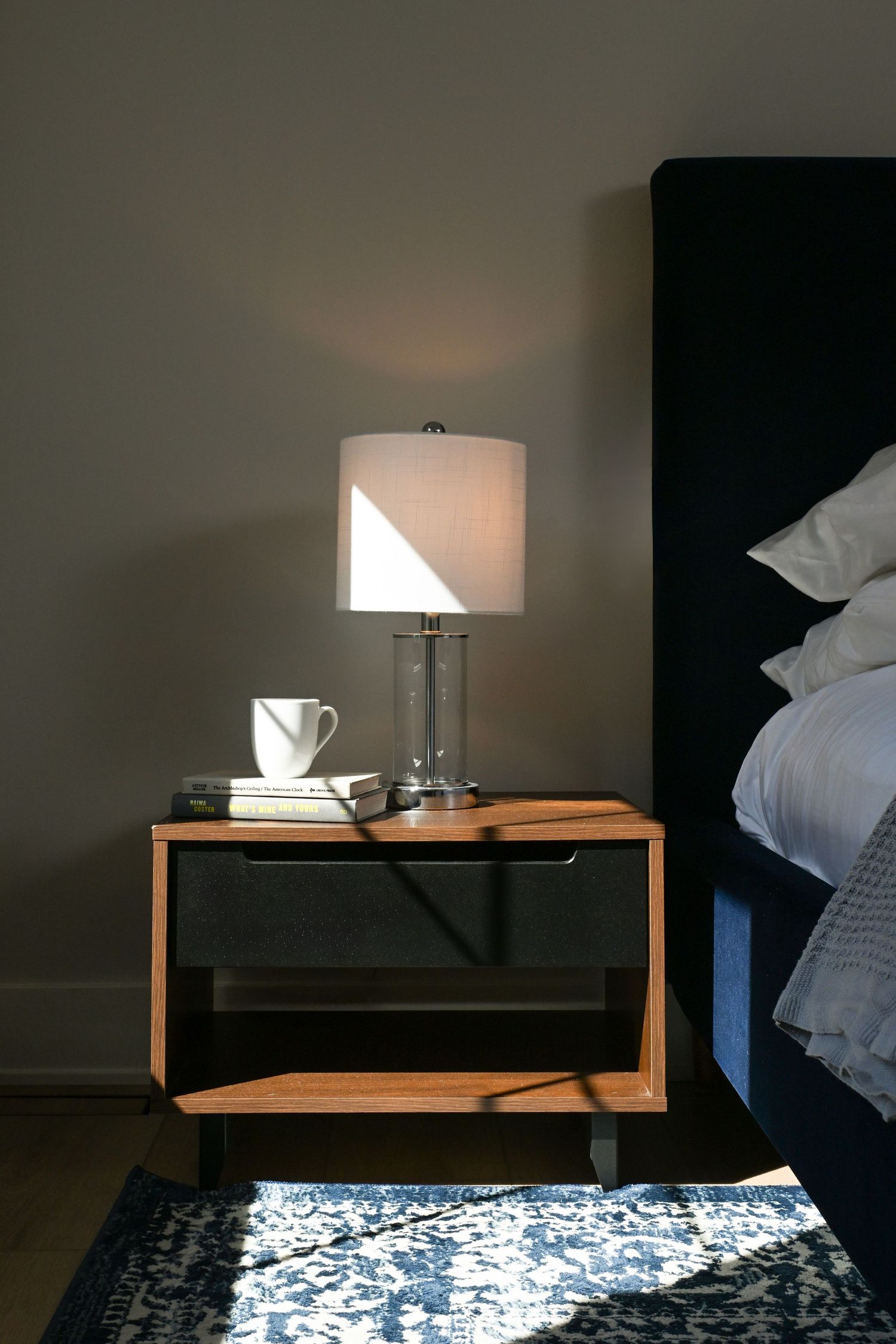 Modern bedroom nightstand with glowing table lamp, coffee mug, and books beside a navy upholstered bed.