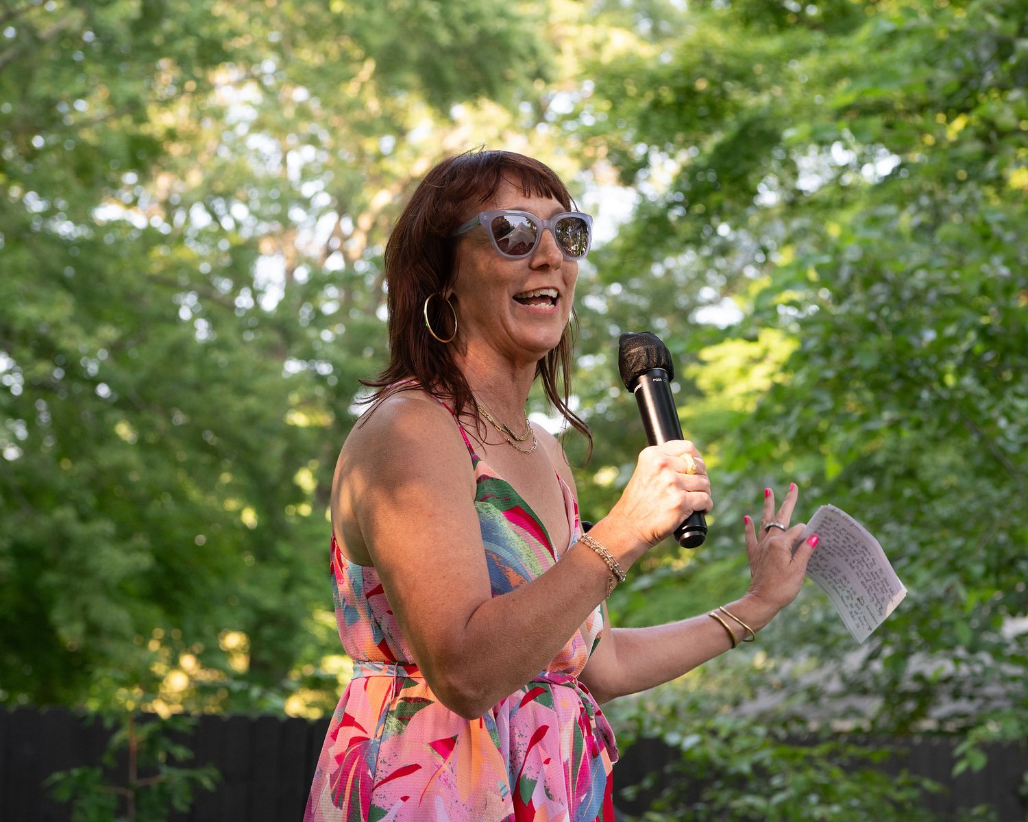 Person in floral dress and sunglasses laughing while holding a drink outdoors on a sunny summer day.