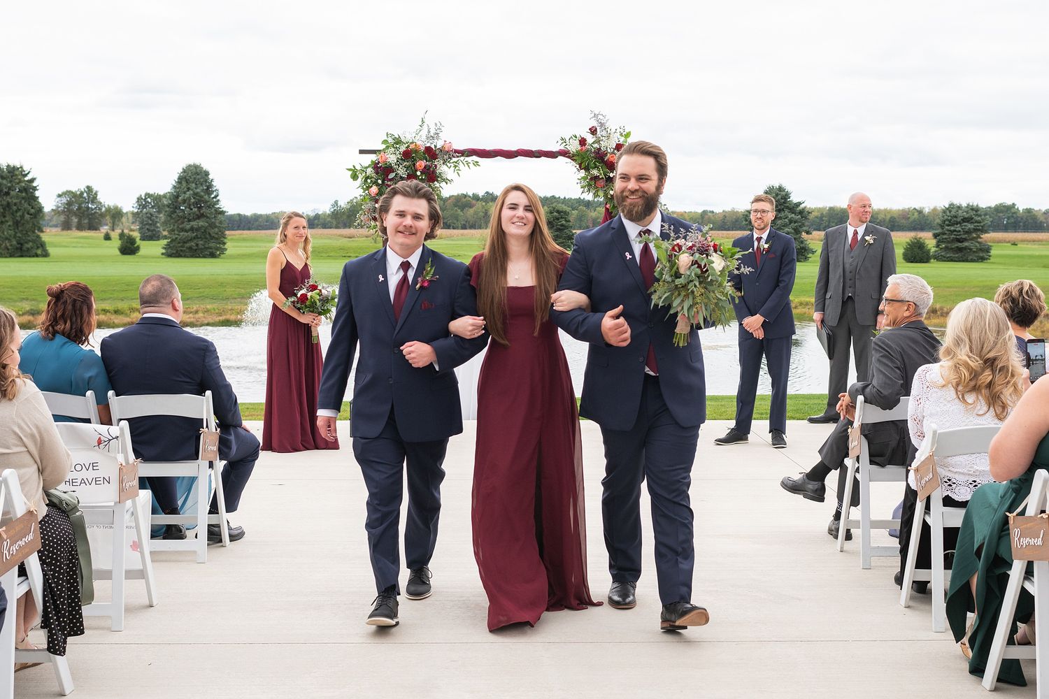 Bride in burgundy gown walks down wedding aisle escorted by two groomsmen in navy suits outdoors.