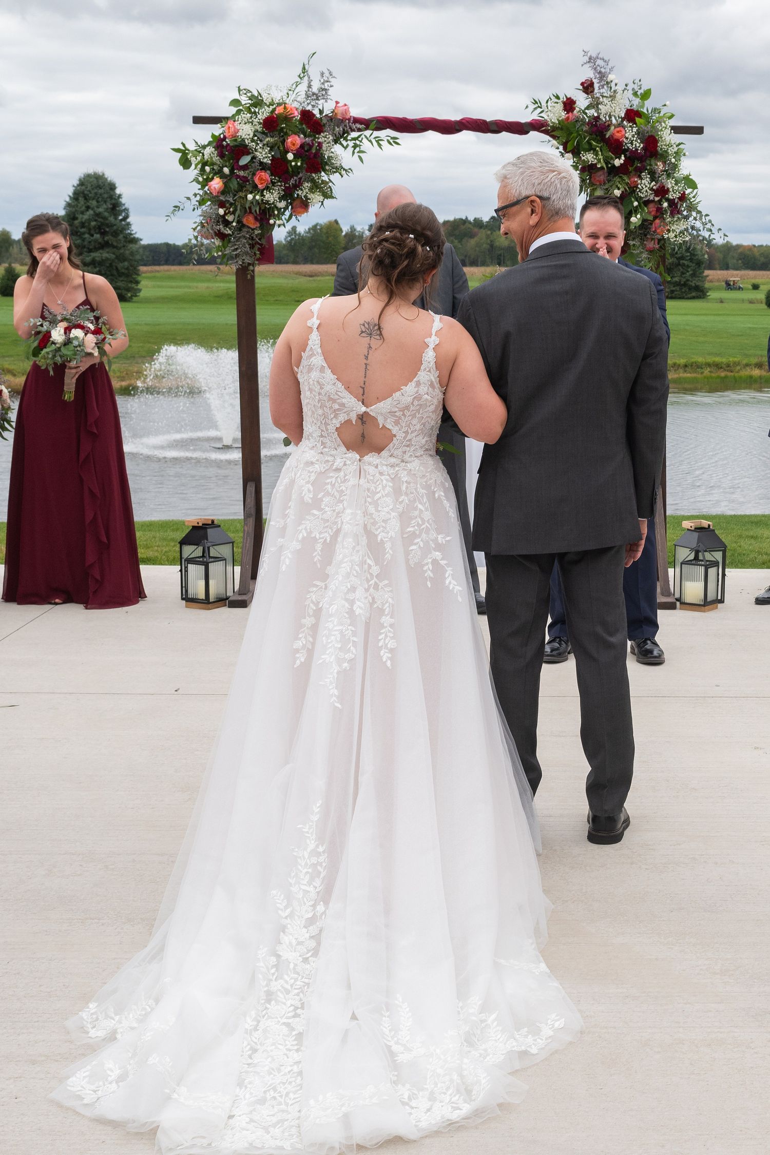 Bride in lace wedding gown stands at floral arch altar with groom, bridesmaid in burgundy dress nearby, outdoor ceremony.