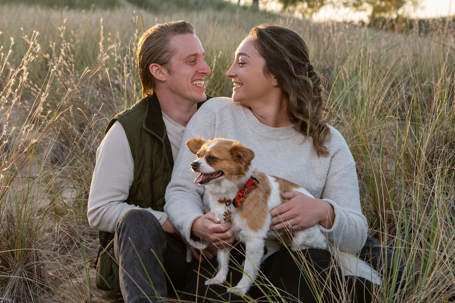 Couple sitting in tall grass field holding a small brown and white dog, smiling at each other during golden hour.