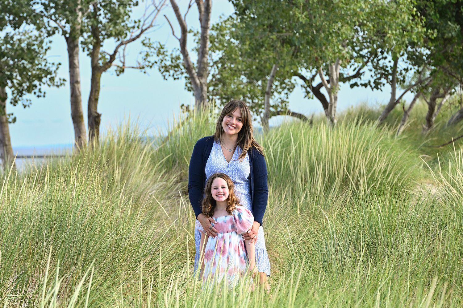A woman and young girl in floral dress pose smiling among tall beach grass with trees and lake in background.
