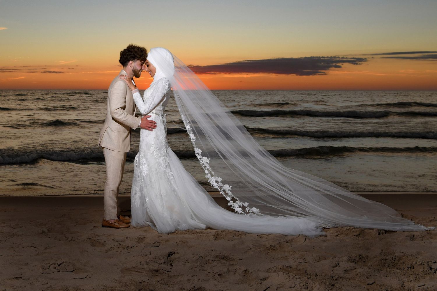 Dramatic sunset beach wedding photo with flowing veil against orange sky.