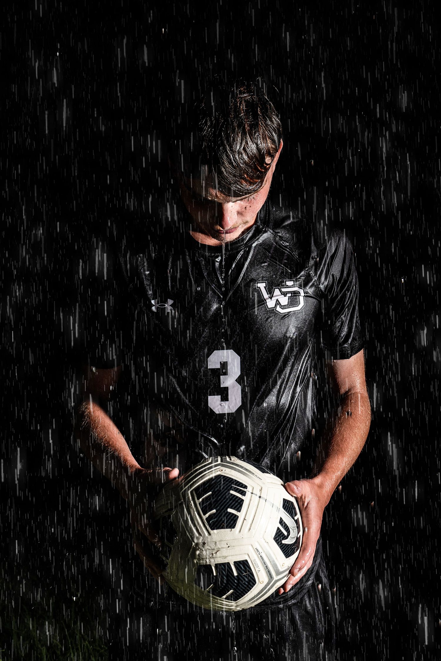 Soccer player in black jersey holding a white ball in dramatic rainy conditions.