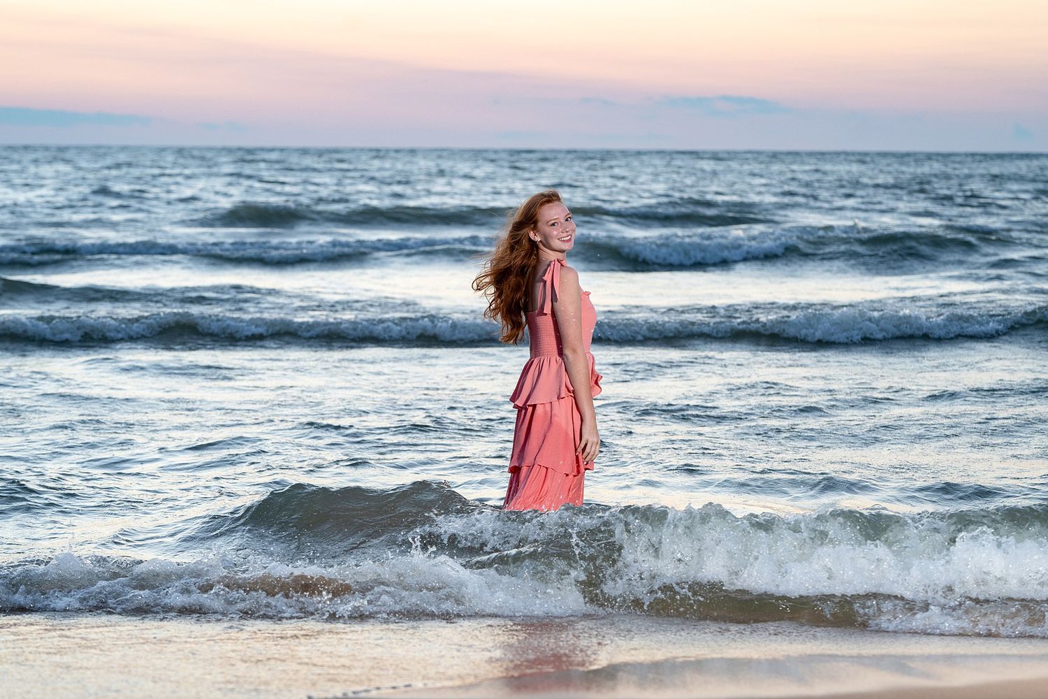 Young woman in pink dress standing in ocean waves at sunset on a beach.
