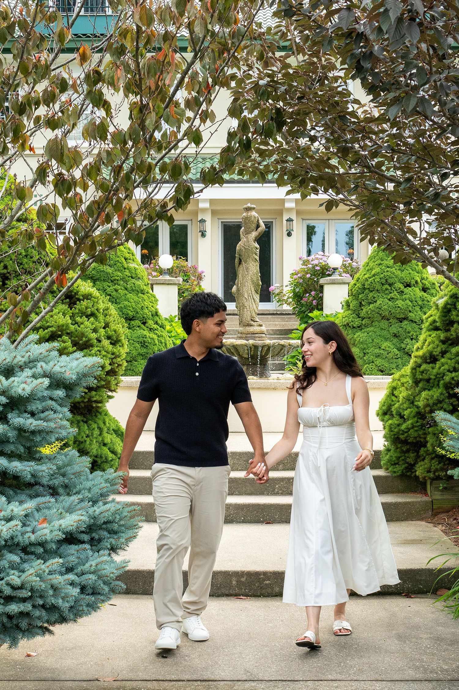 Couple holding hands walking through a lush garden with a fountain and elegant white building in the background.