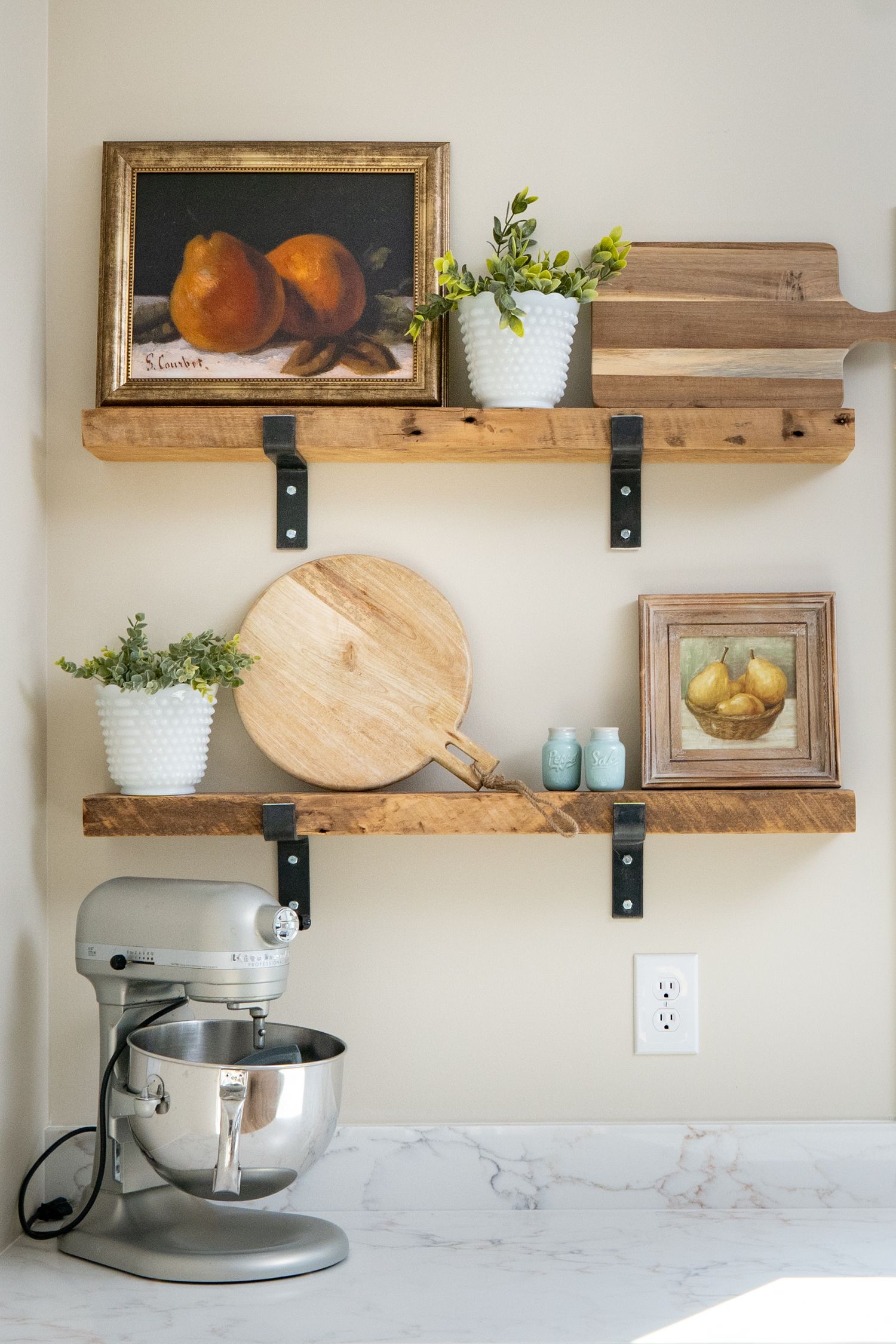 Rustic wooden kitchen shelves with fruit paintings, white planters, a cutting board, and a stand mixer on marble countertop.