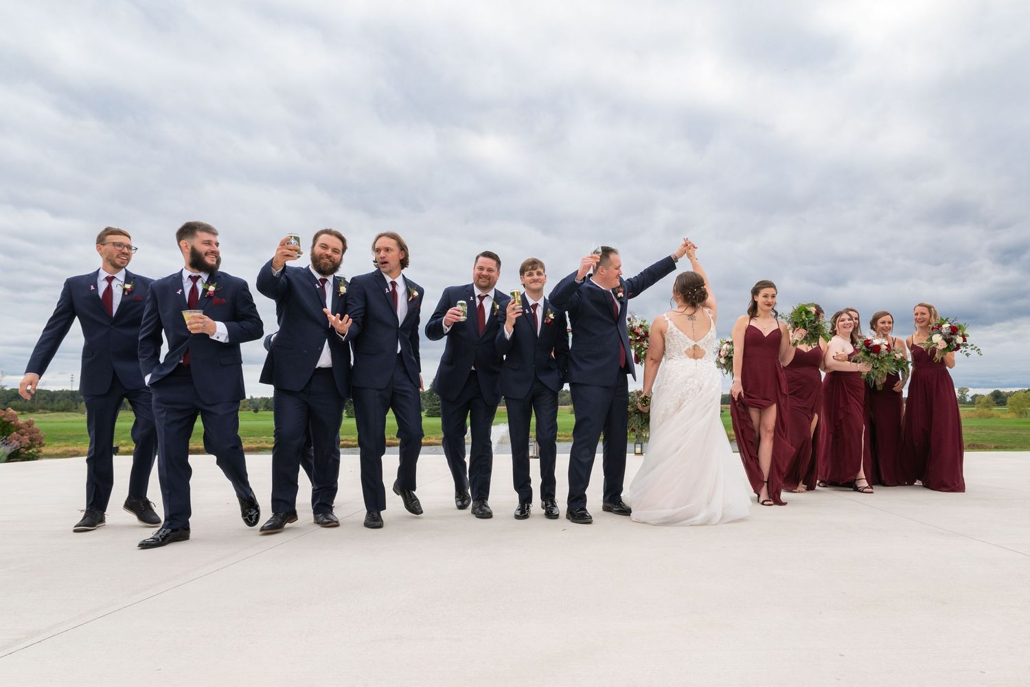 Wedding party celebrating outdoors, groomsmen in navy suits and bridesmaids in burgundy dresses cheering under cloudy skies.