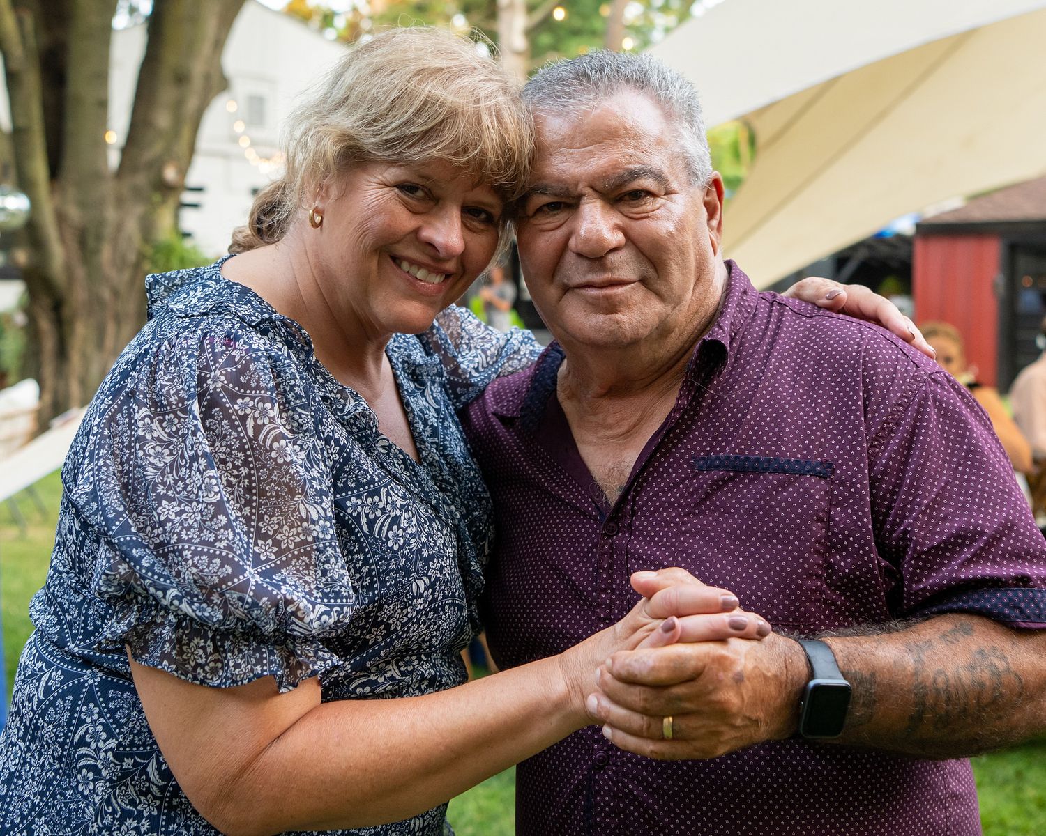 A couple holds hands and smiles together at an outdoor event wearing casual attire.
