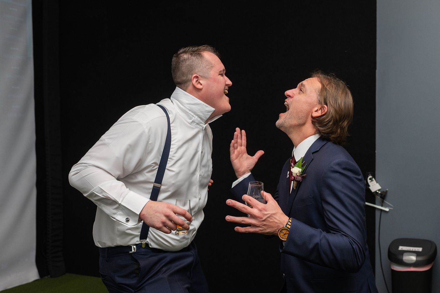 Two men in formal suits laugh and gesture animatedly at a wedding reception photo booth.