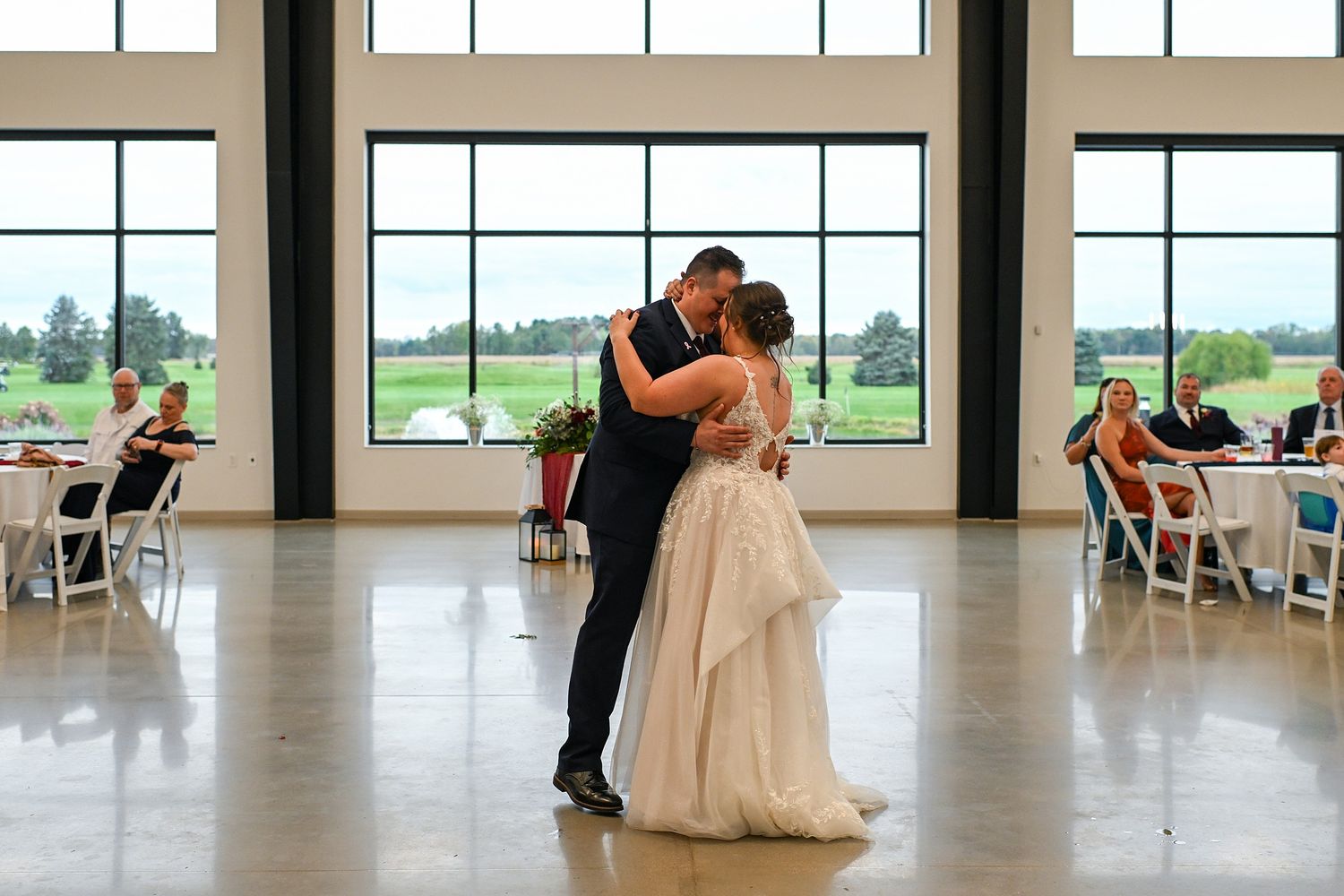 Bride and groom share their first dance in a bright modern venue with large windows and seated guests in the background.