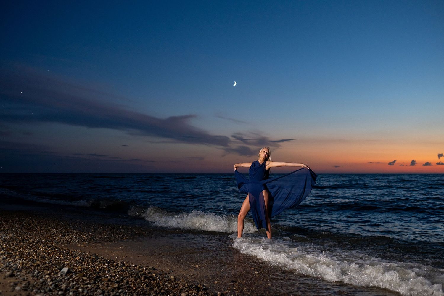 Dancer performs with flowing dress against twilight sky with moon visible over ocean waves.