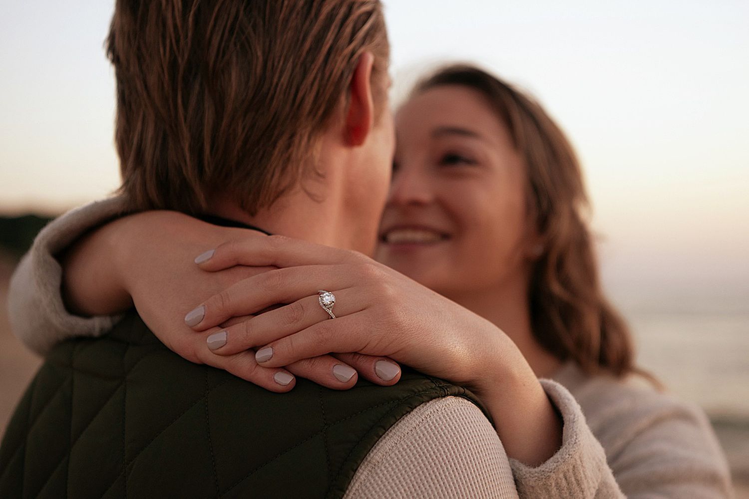 Couple embracing at sunset, woman smiling and showing off a diamond engagement ring on her hand.