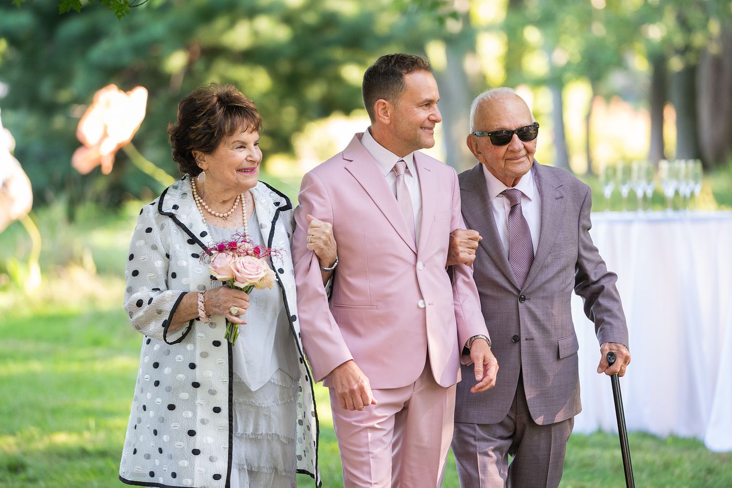 Three people walking together at an outdoor event, with one wearing a pink suit and others in polka dots and gray attire.