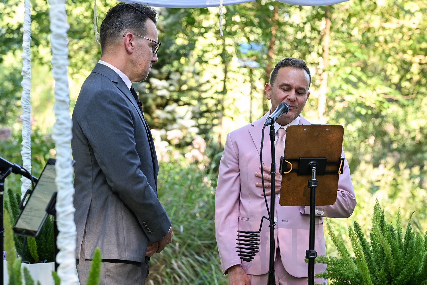 Two people stand at an outdoor ceremony, one in a gray suit and one in a pink suit reading from a clipboard in a garden setting.