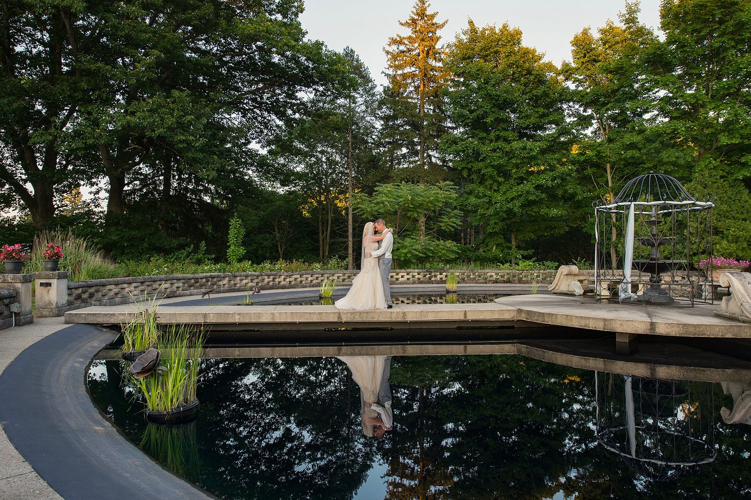 Bride and groom embrace on a bridge over a reflective pond in a lush garden wedding venue at golden hour.