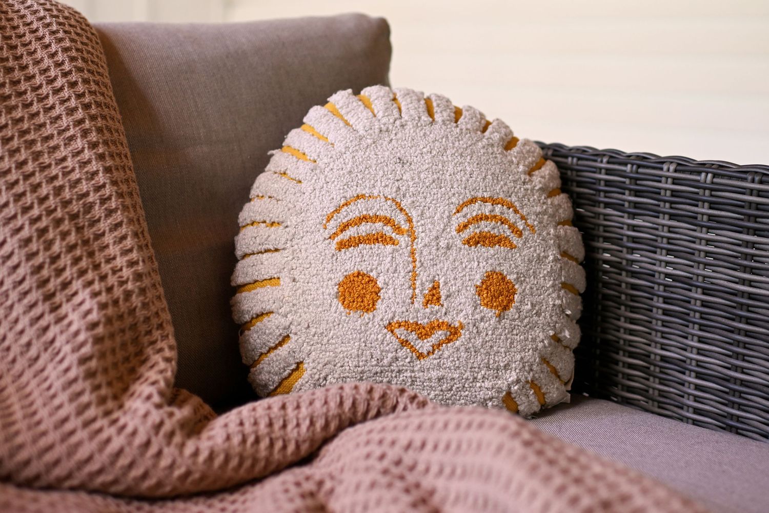 Round decorative sun face pillow with golden embroidery on a gray wicker sofa with pink knit blanket.