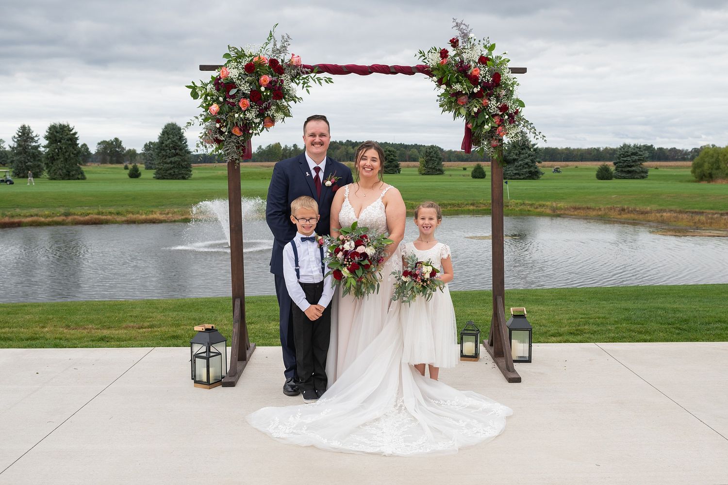 Bride and groom pose with young ring bearer and flower girl under floral arch by pond at outdoor wedding ceremony.