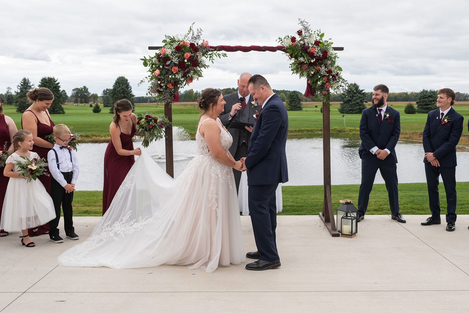 Bride and groom exchange vows under a floral arch by a pond with wedding party beside them.