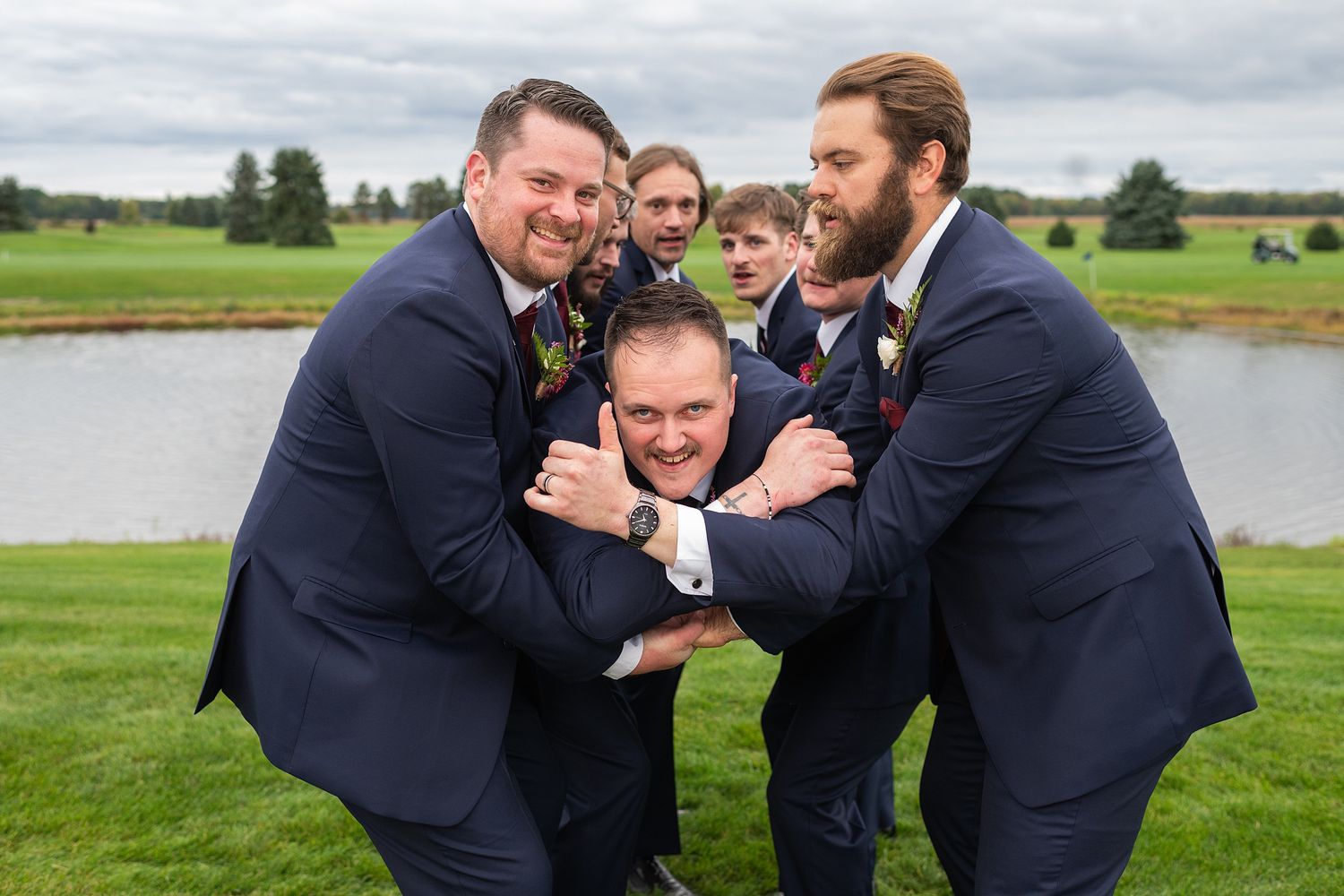 Groomsmen in navy suits playfully lifting the groom outdoors near a pond at a golf course wedding.