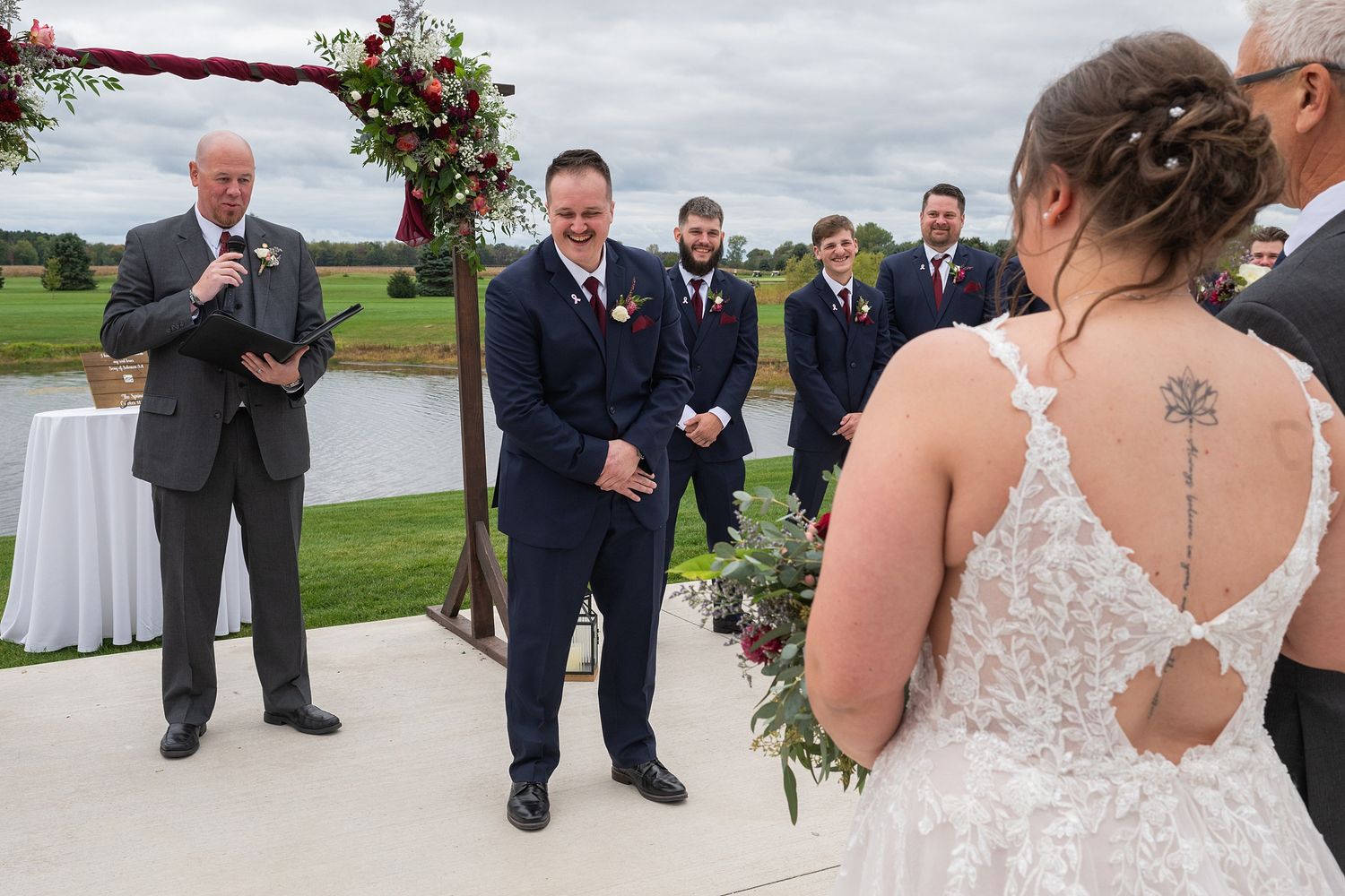 Groom smiles emotionally as bride approaches the outdoor wedding ceremony altar by a pond.
