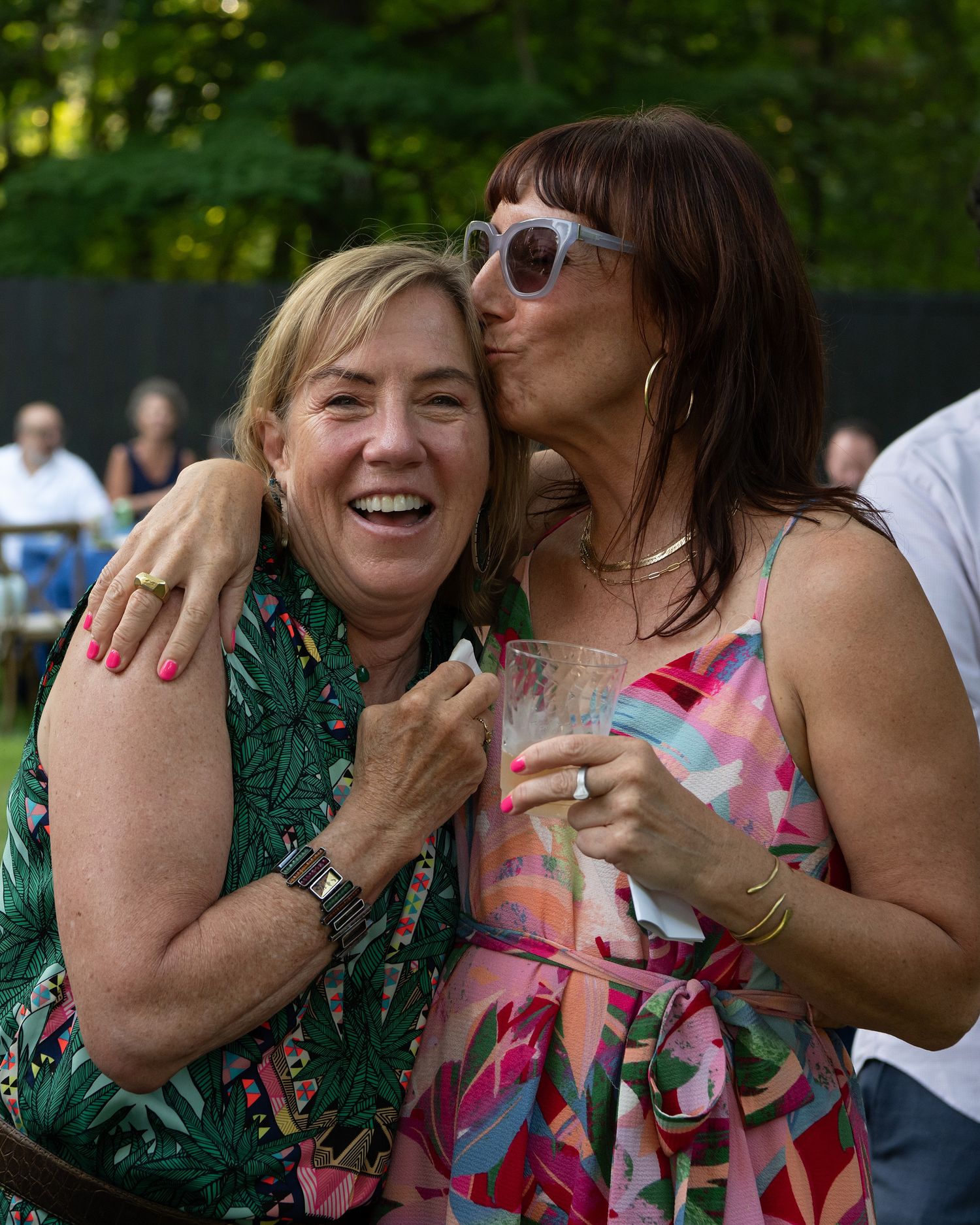 Two people laughing and embracing at an outdoor gathering with lush green foliage in the background.