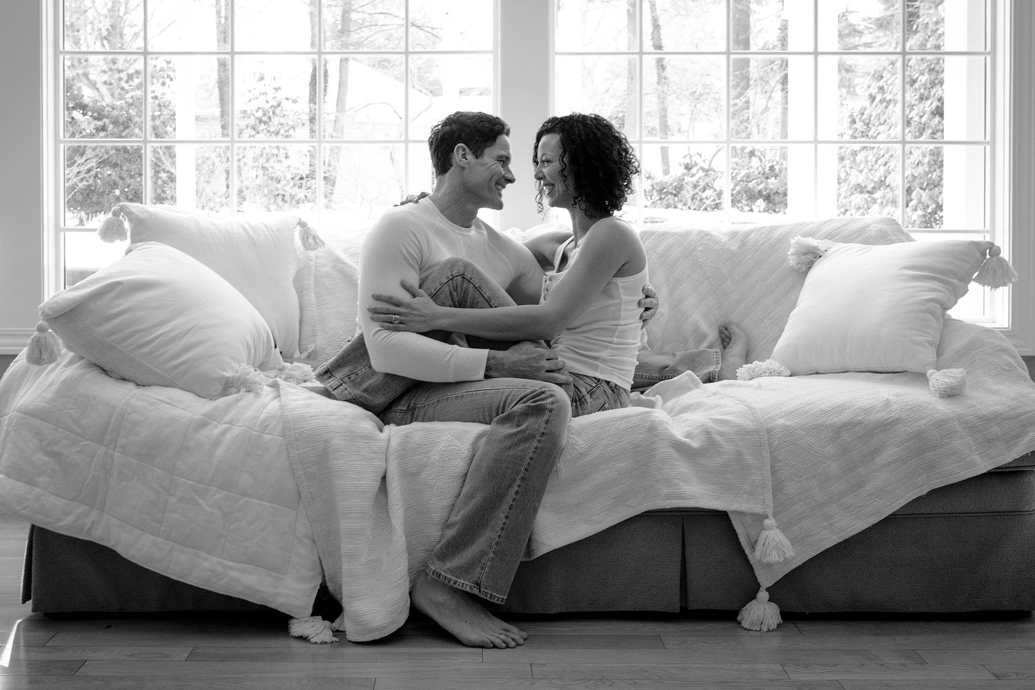 Couple sitting intimately on a cozy sofa, gazing at each other in a bright, sunlit living room, black and white photo.
