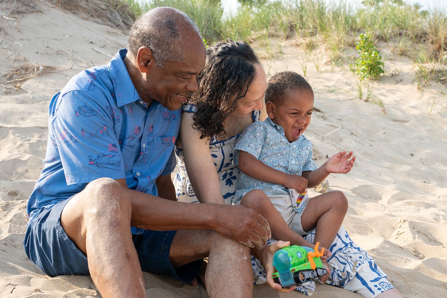 Grandparents and young child sitting together on sandy beach, laughing and playing outdoors on a sunny day.