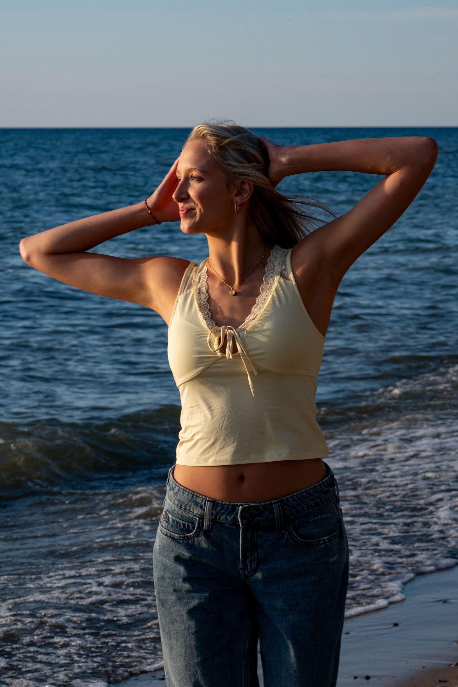 A person in jeans and tank top poses against ocean waves on a rocky shoreline.