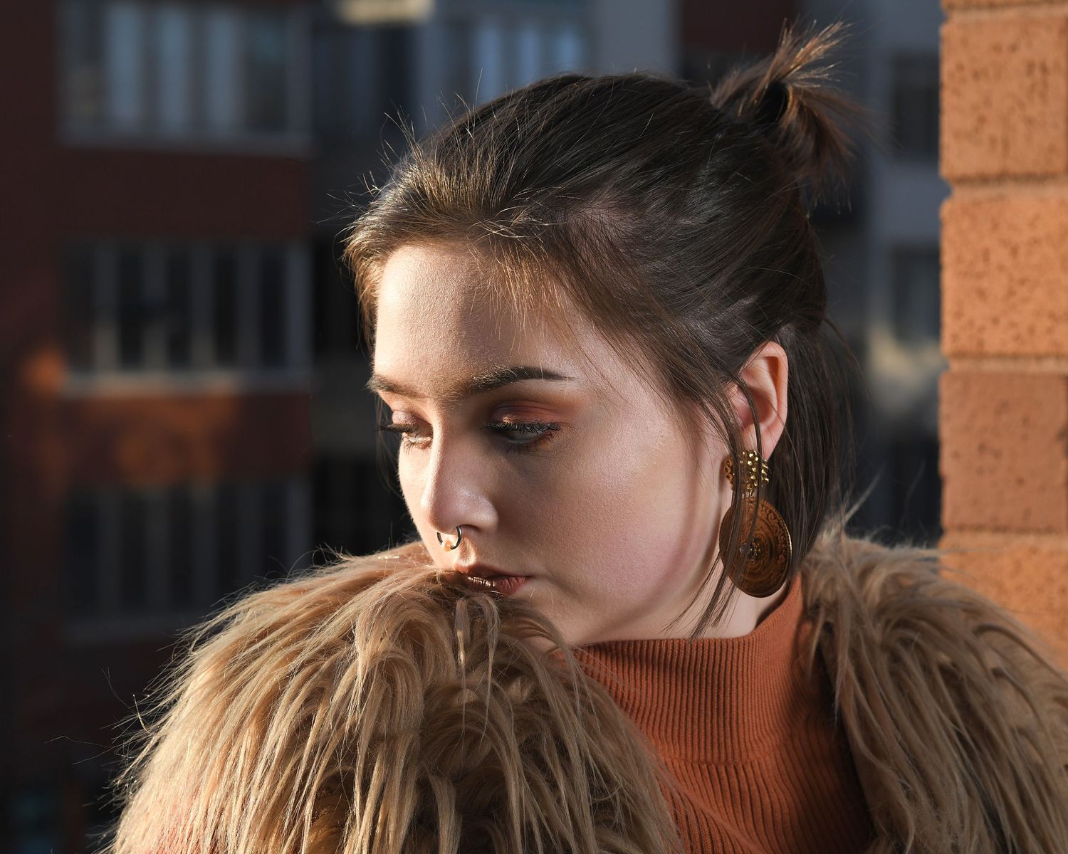 Moody portrait with fur coat and gold hoop earrings against brick wall.