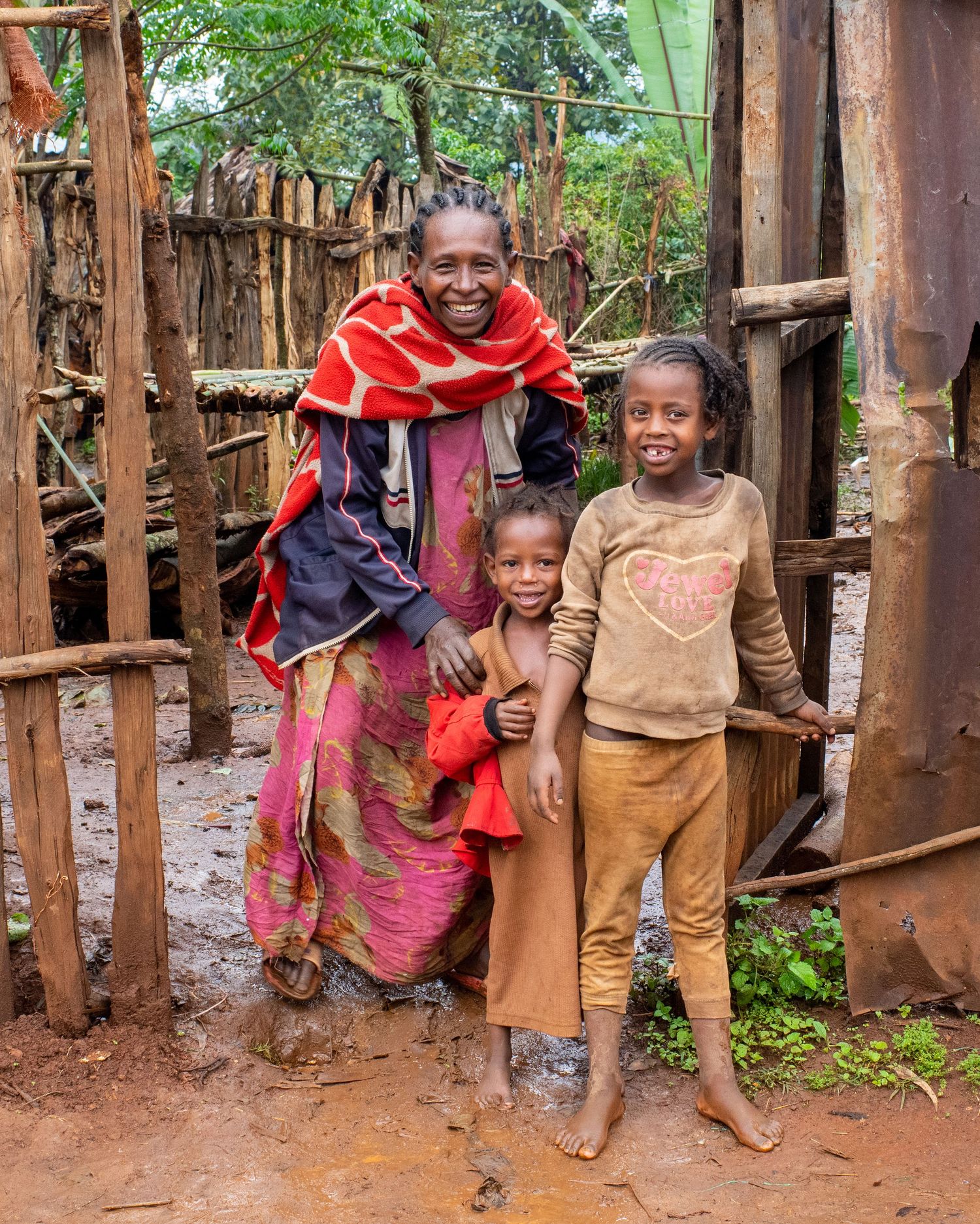 A family group poses together outside their home with wooden fence and mud walls.