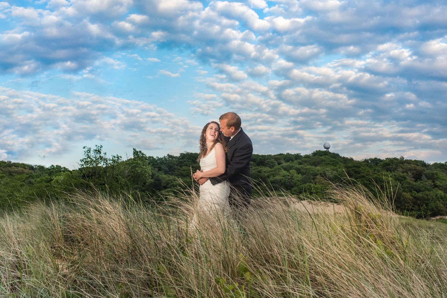 Couple embraces in tall grass field under cloudy blue sky.