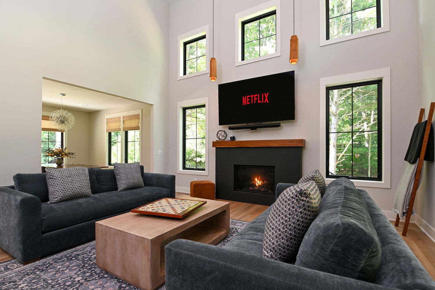 Modern living room with gray sofas, wood fireplace, Netflix TV, high ceilings, and large windows overlooking trees.