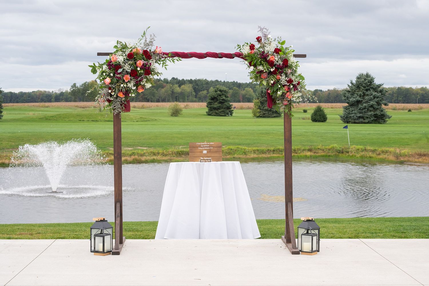Elegant outdoor wedding arch adorned with red and white florals overlooking a serene pond with fountain.