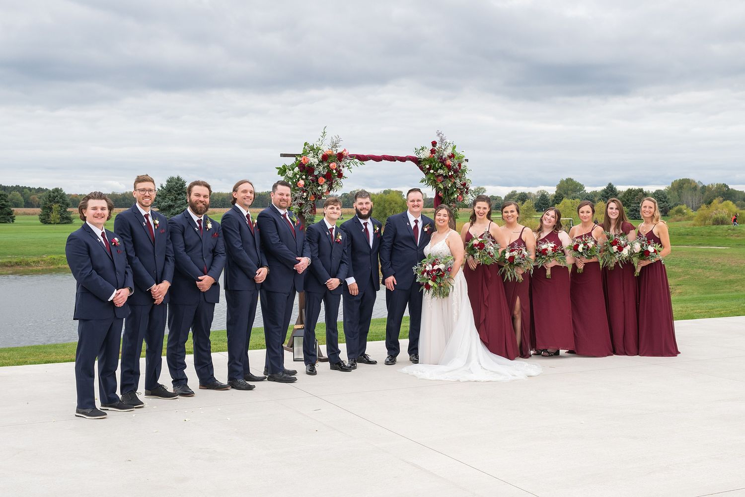 Wedding party poses outdoors with groomsmen in navy suits and bridesmaids in burgundy gowns.