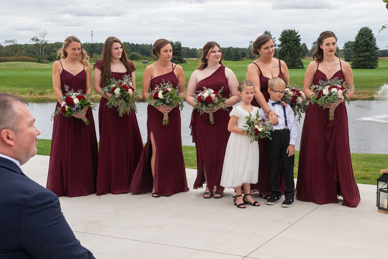 Six bridesmaids in burgundy gowns hold bouquets with flower girl and ring bearer at waterfront venue.