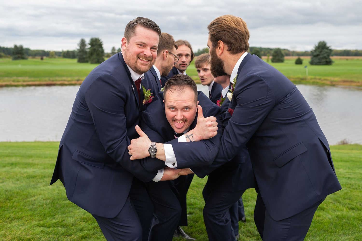 Groomsmen in navy suits playfully tackle the groom outdoors near a pond at a wedding.