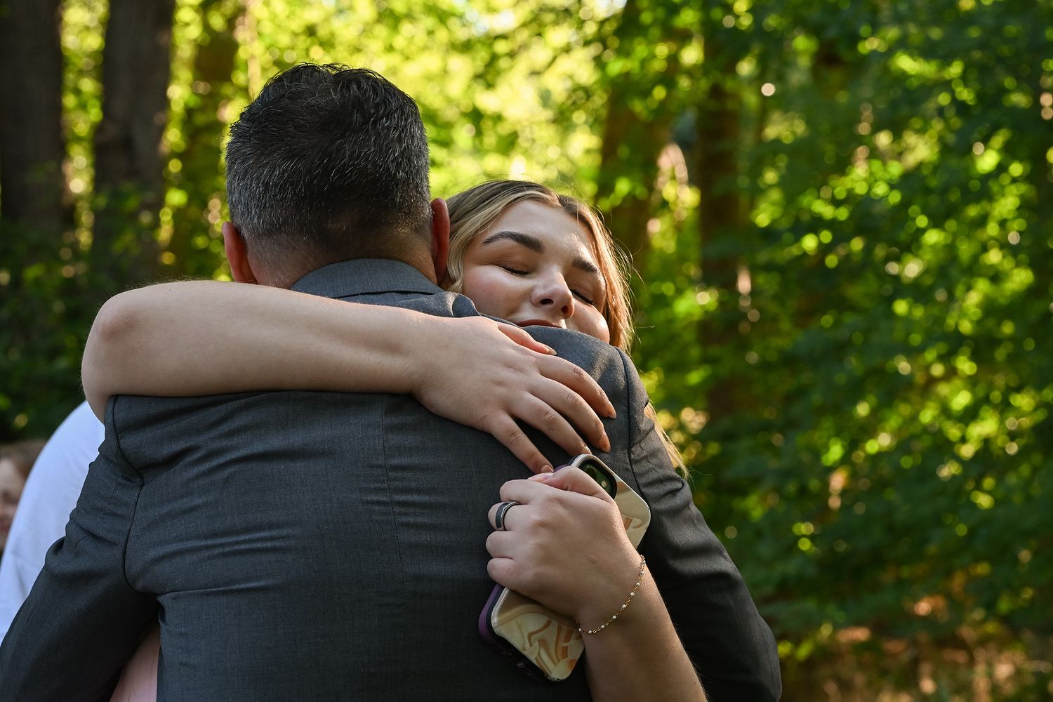 A couple embraces in a sunlit forest setting during what appears to be an intimate outdoor photoshoot.