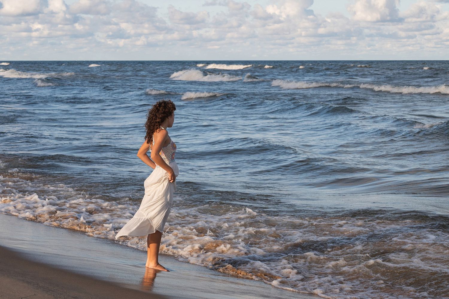 Person in a white dress walks along the shoreline as waves roll in.
