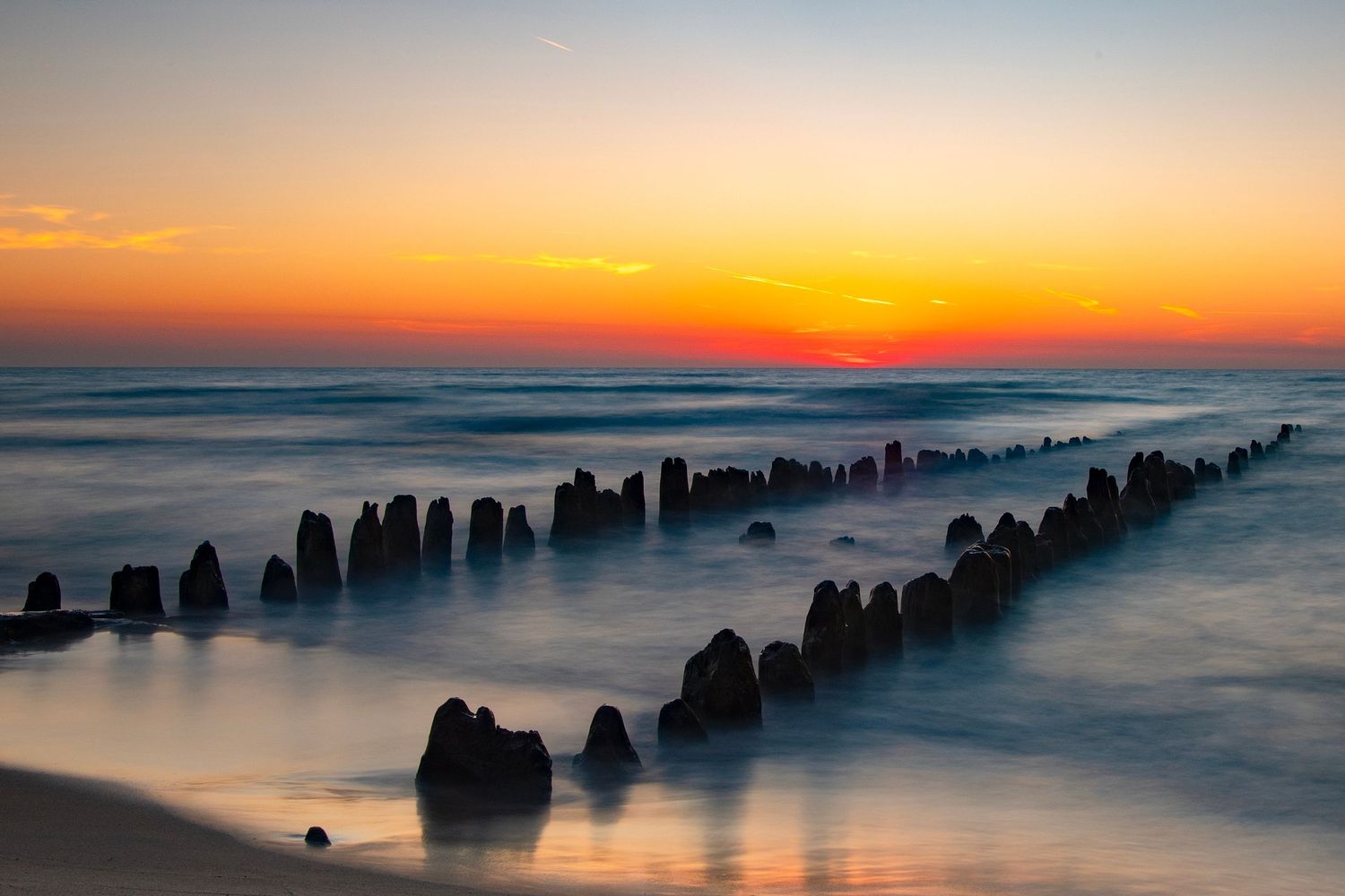 Long wooden posts emerge from misty ocean waters during a vibrant orange sunset.