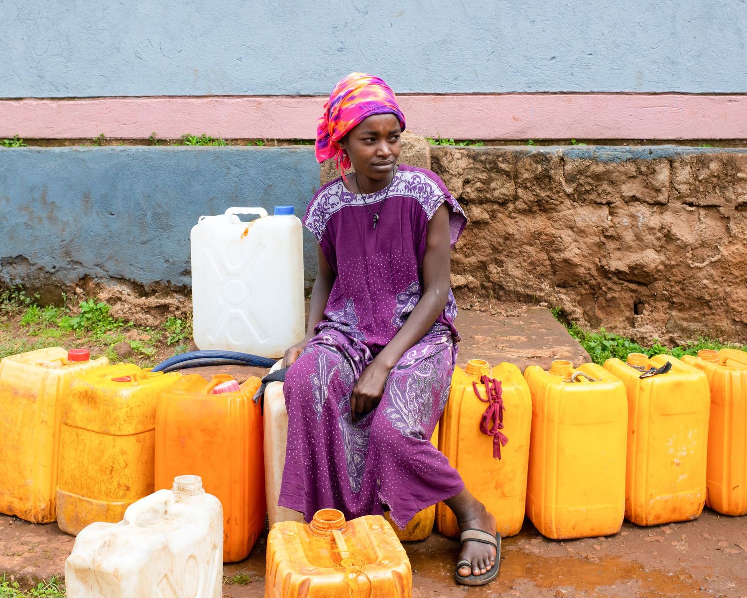 A woman in purple dress sits among yellow water containers outside a building.