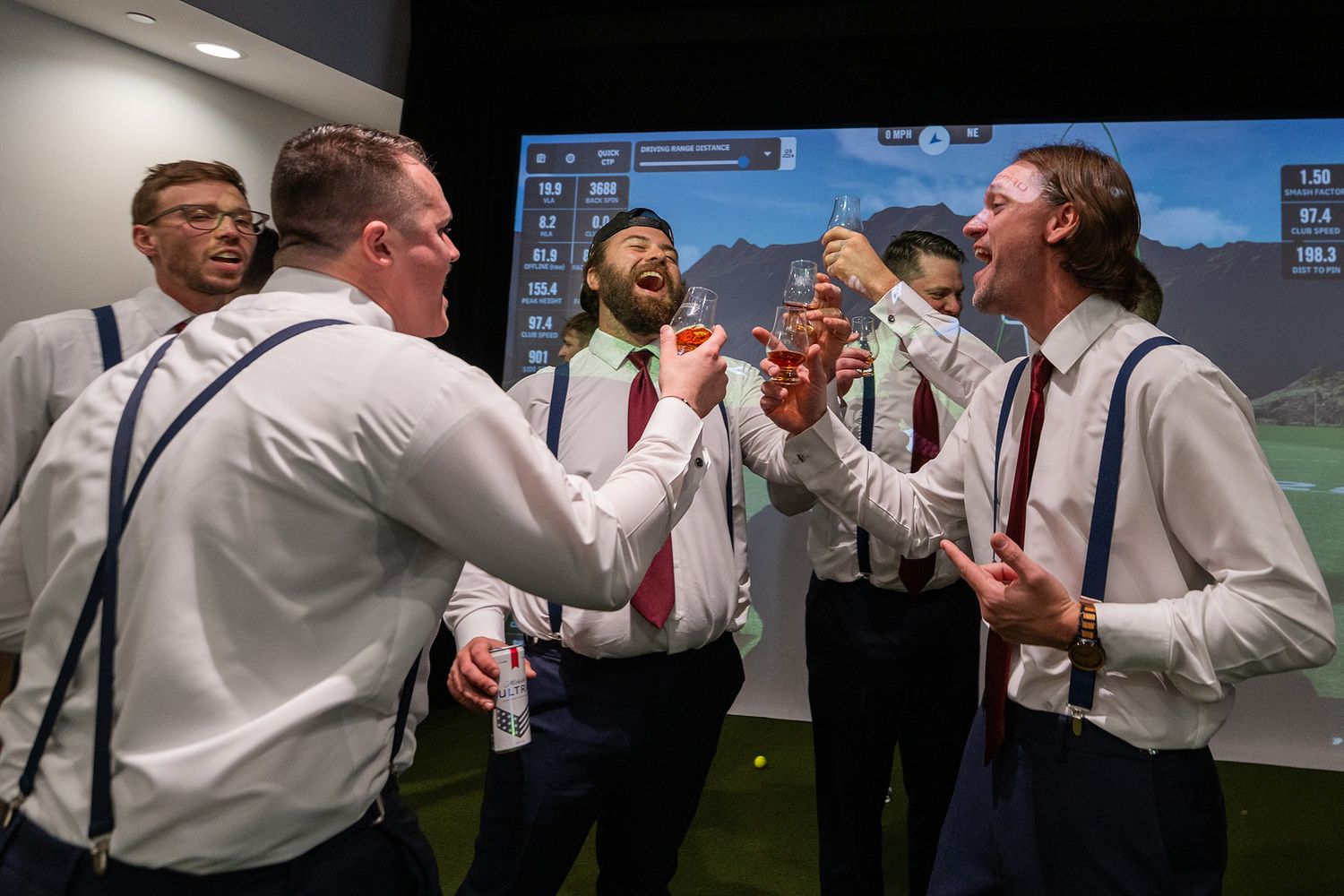 Groomsmen in white shirts and suspenders celebrate joyfully at an indoor golf simulator during wedding festivities.