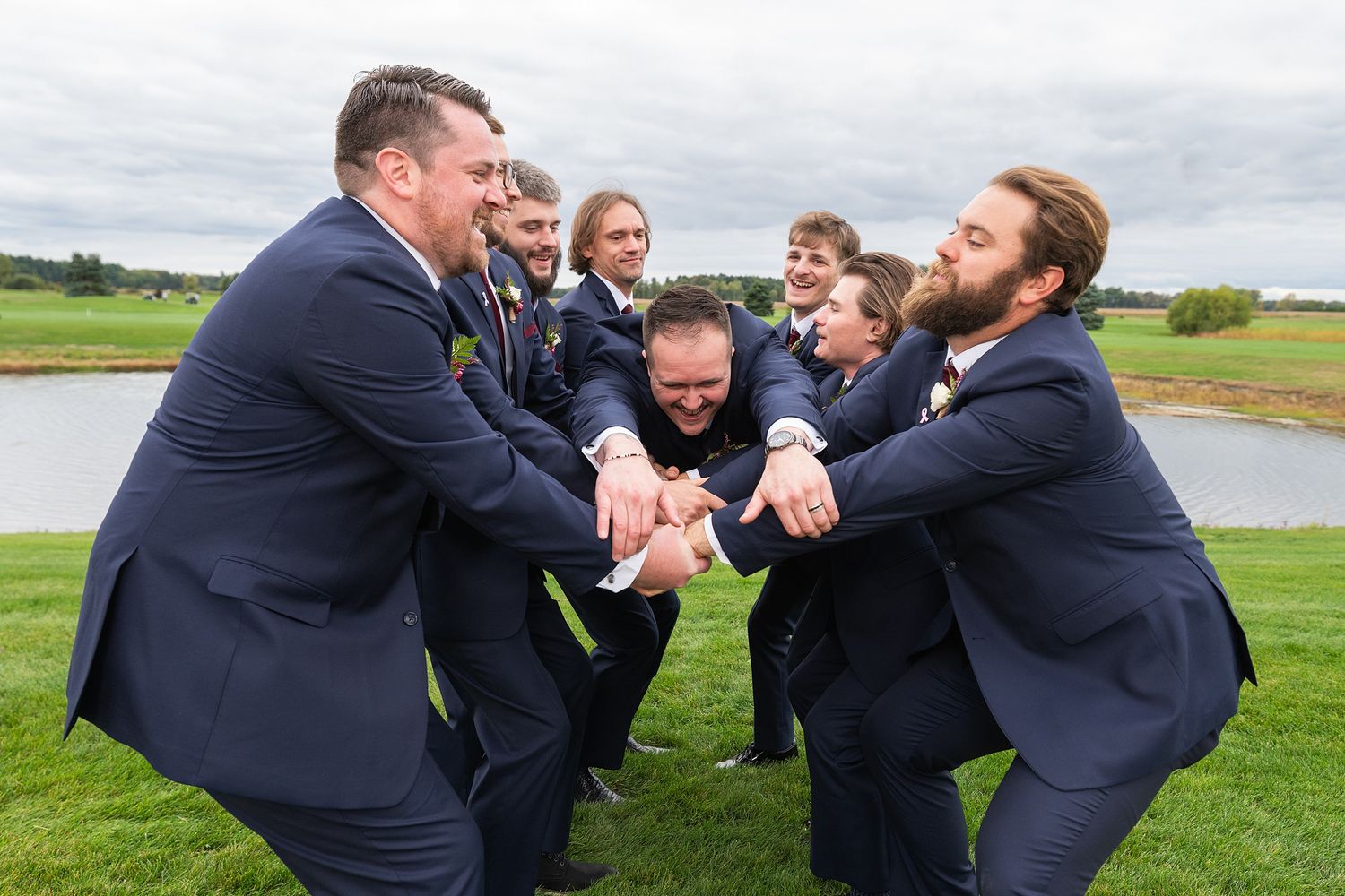 Groomsmen in navy suits laughing and pulling each other in a fun outdoor wedding photo near a pond.
