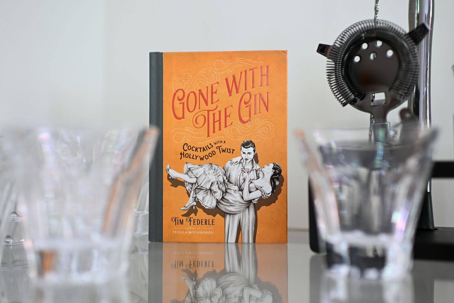 Gone with the Gin cocktail recipe book displayed beside bar tools and glassware on a reflective surface.